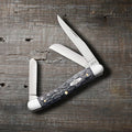  Case Pocket Worn Crandall Jig Gray Bone Medium Stockman pocket knife with Clip, Sheepfoot, and Spey blades open, on a rustic wood surface