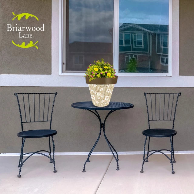 Medium tan floral planter filled with flowers on a black patio table between two matching chairs.