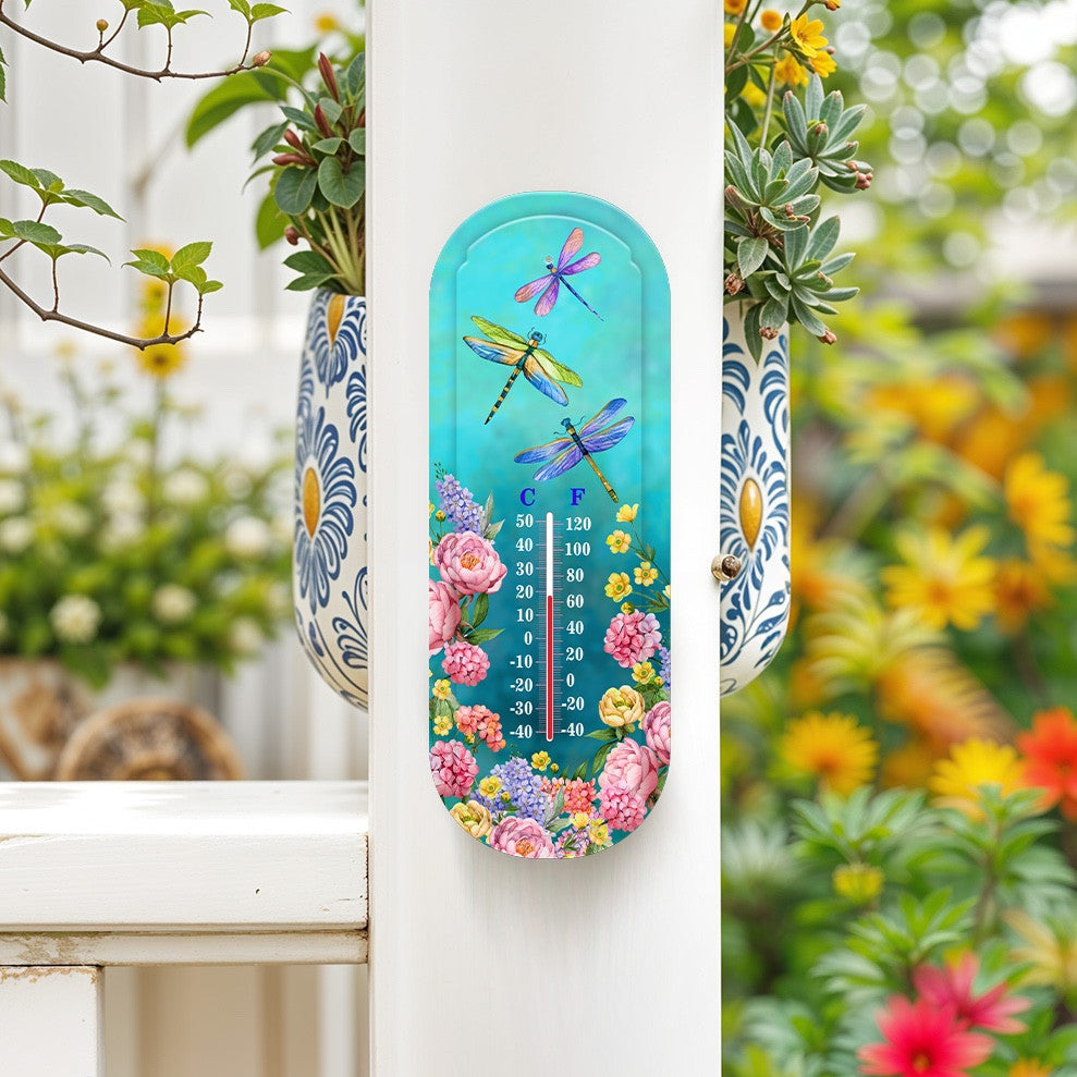 Turquoise dragonfly and floral thermometer mounted on a white porch post beside flowering plants.