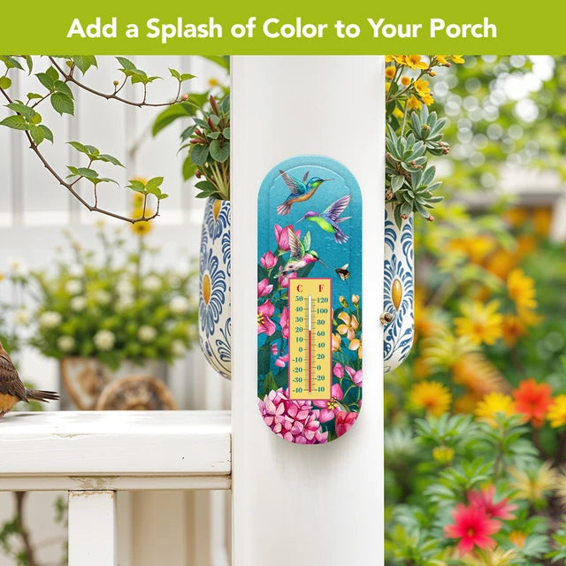 Turquoise hummingbird and floral thermometer mounted on a white porch post beside flowering plants.