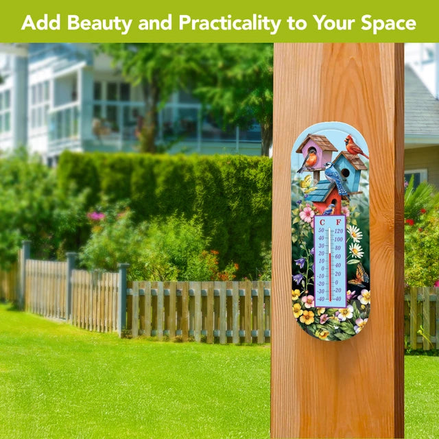Birdhouse and floral metal thermometer mounted on a wooden post overlooking a green lawn and picket fence.