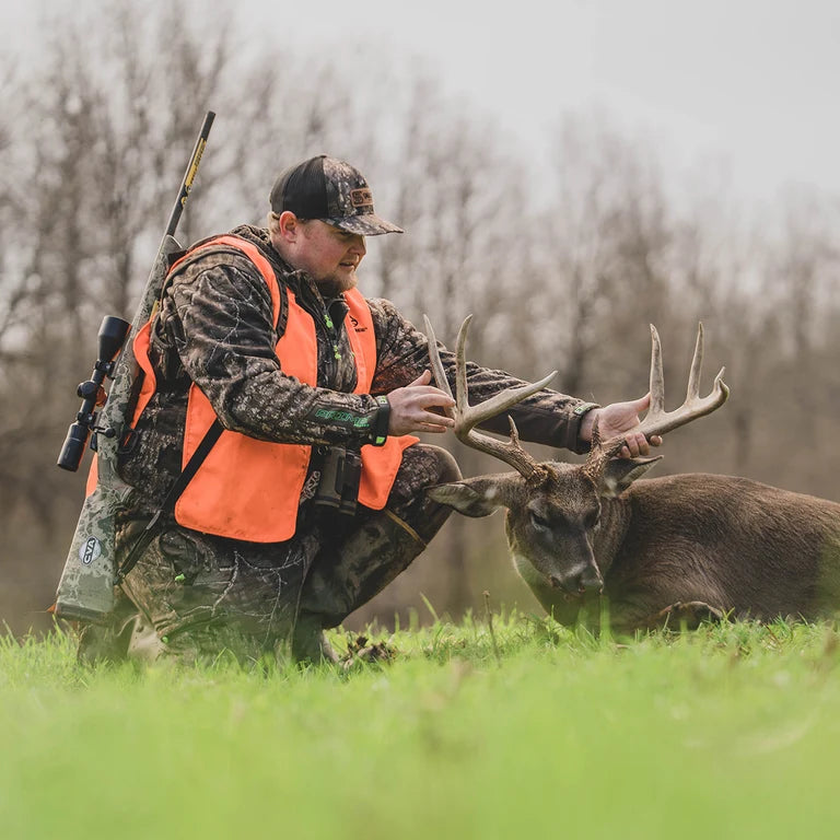 Hunter wearing camouflage and an orange vest kneeling beside a harvested buck in a green field.