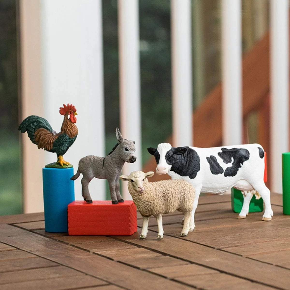 Schleich donkey, rooster, sheep, and cow figures arranged with colorful blocks on a wooden table.