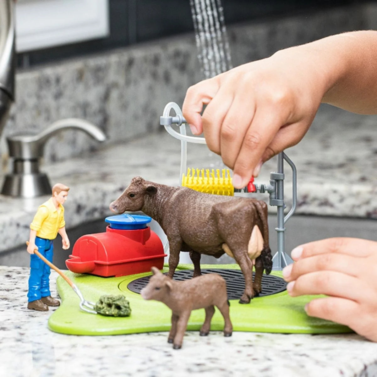 Hands operating the Schleich Happy Cow Wash station with a cow beneath the yellow brush and a calf nearby.