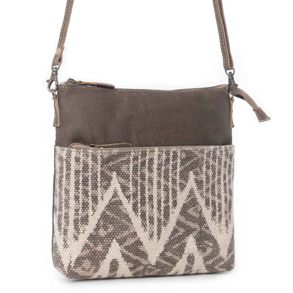 Angled view of Myra Coffee Canvas Crossbody Bag with genuine leather and neutral patterned rug detailing
