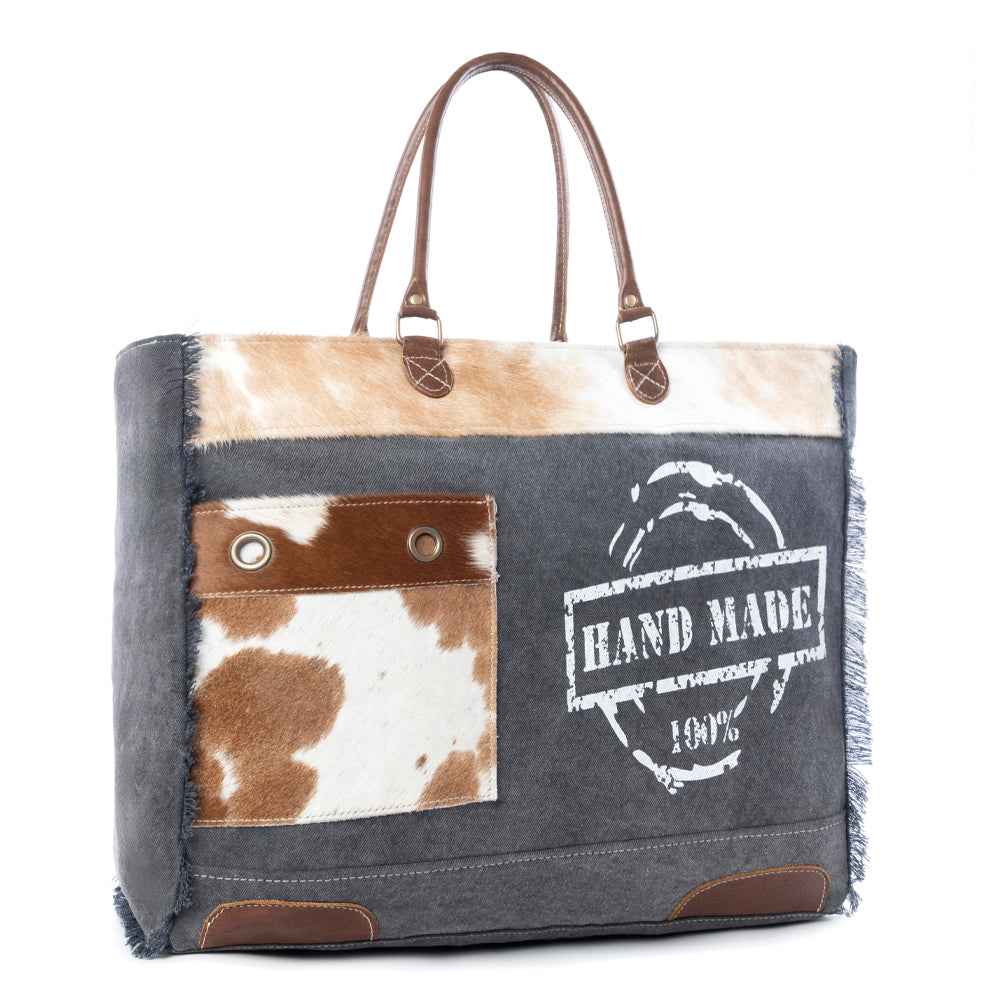 Angled view of Myra Dakota Trail Weekender Bag with hair-on hide accents, dark canvas, leather handles and fringe details