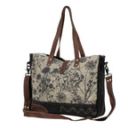 Angled view of Myra Mystique messenger bag featuring floral canvas, genuine leather details and dual carrying straps.