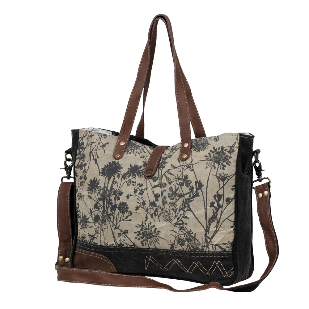 Angled view of Myra Mystique messenger bag featuring floral canvas, genuine leather details and dual carrying straps.