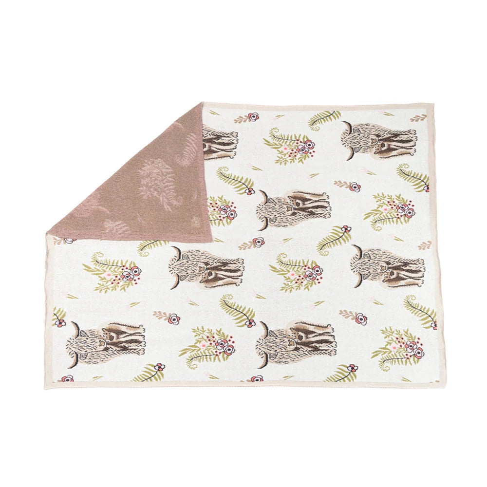 Cream throw blanket with Highland cattle, flowers, and green foliage, with a corner folded back.