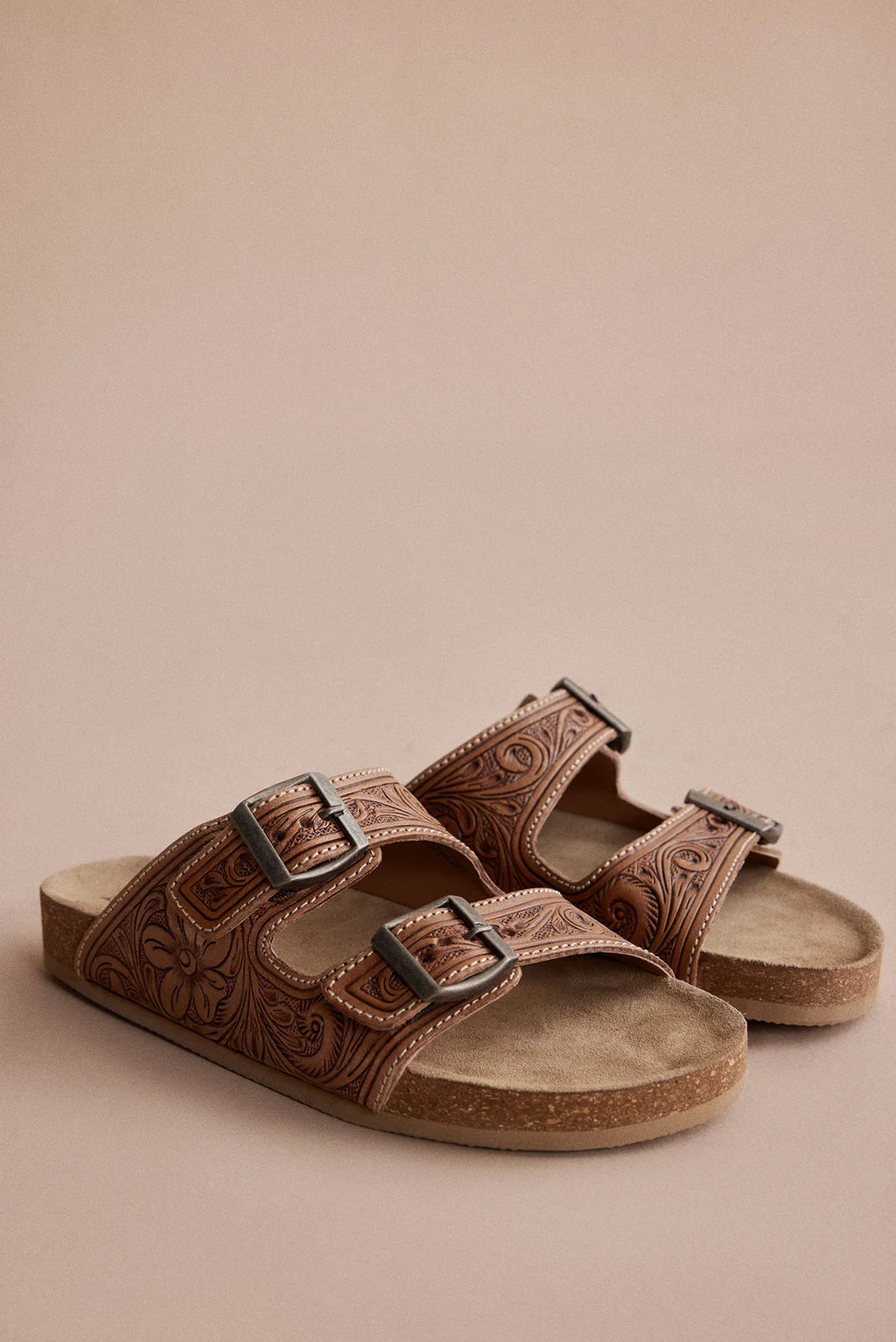 Pair of Myra Darla Trail caramel leather sandals with floral tooling and double-buckle straps on a tan background.