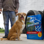 A tan dog sitting beside a man on concrete, next to a blue Sportmix High Energy 26/18 bag leaning against a large tire in front of a grain bin.